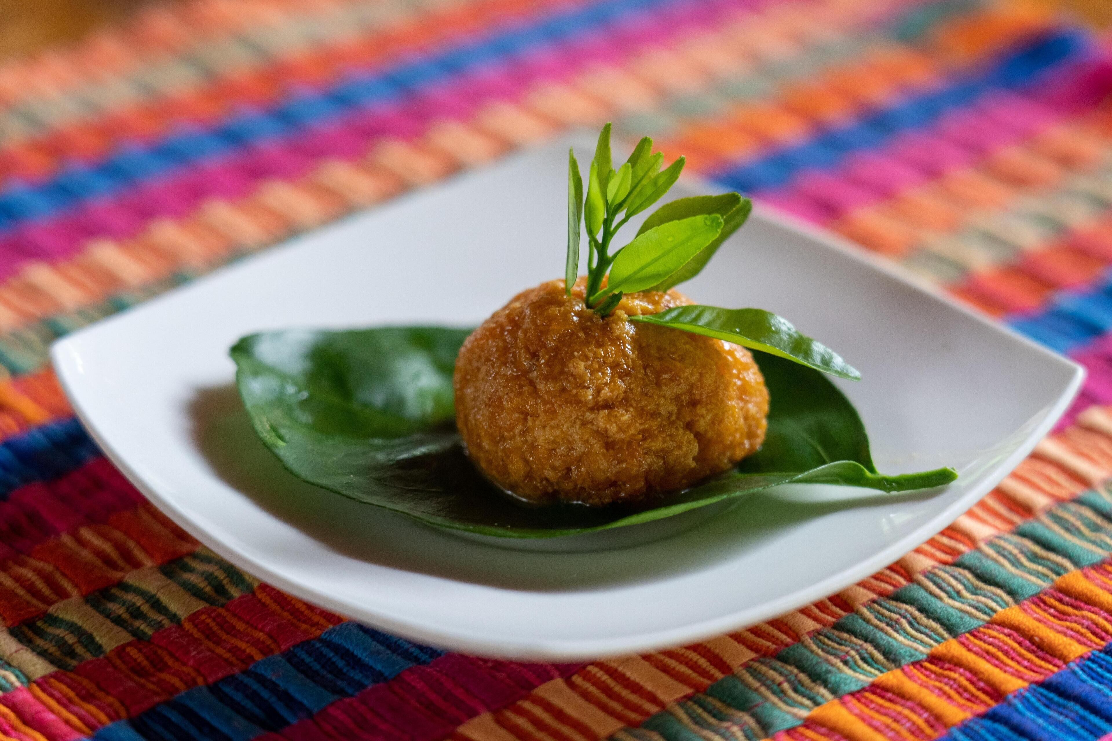 A round, golden-brown food item on a white plate with a green leaf garnish, placed on a colorful woven tablecloth.
