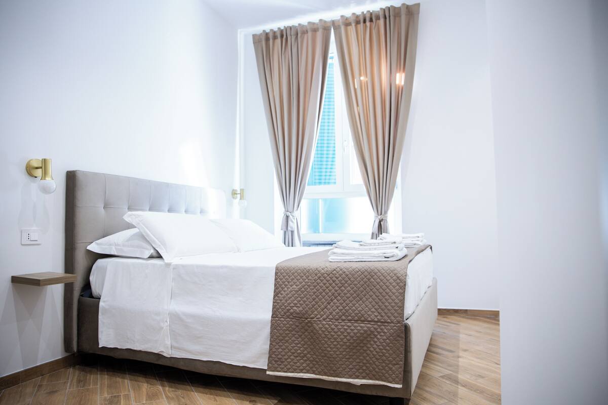 Ein ordentlich bezogenes Bett mit weißer und brauner Bettdecke, ein Nachttisch mit Lampe sowie ein Fenster mit Vorhängen.