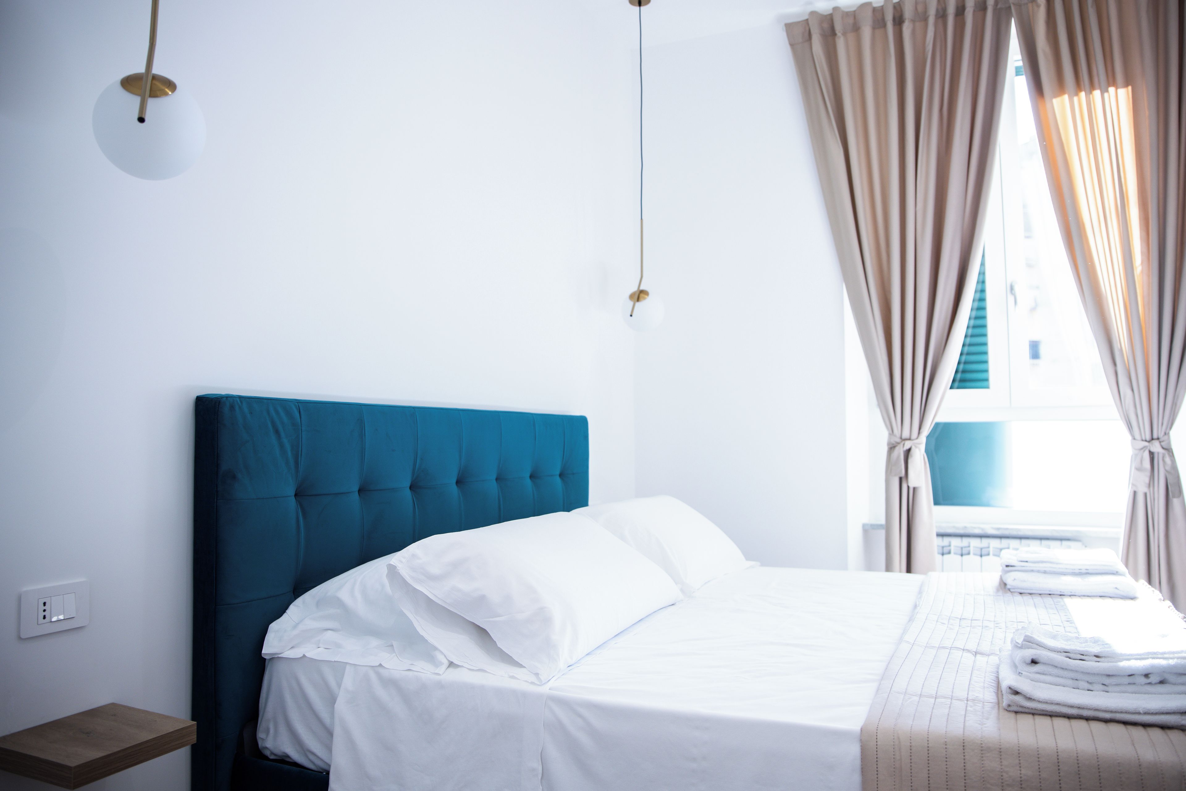 Habitación de hotel con cama, mesita de noche, ventana con cortinas y toma de corriente.