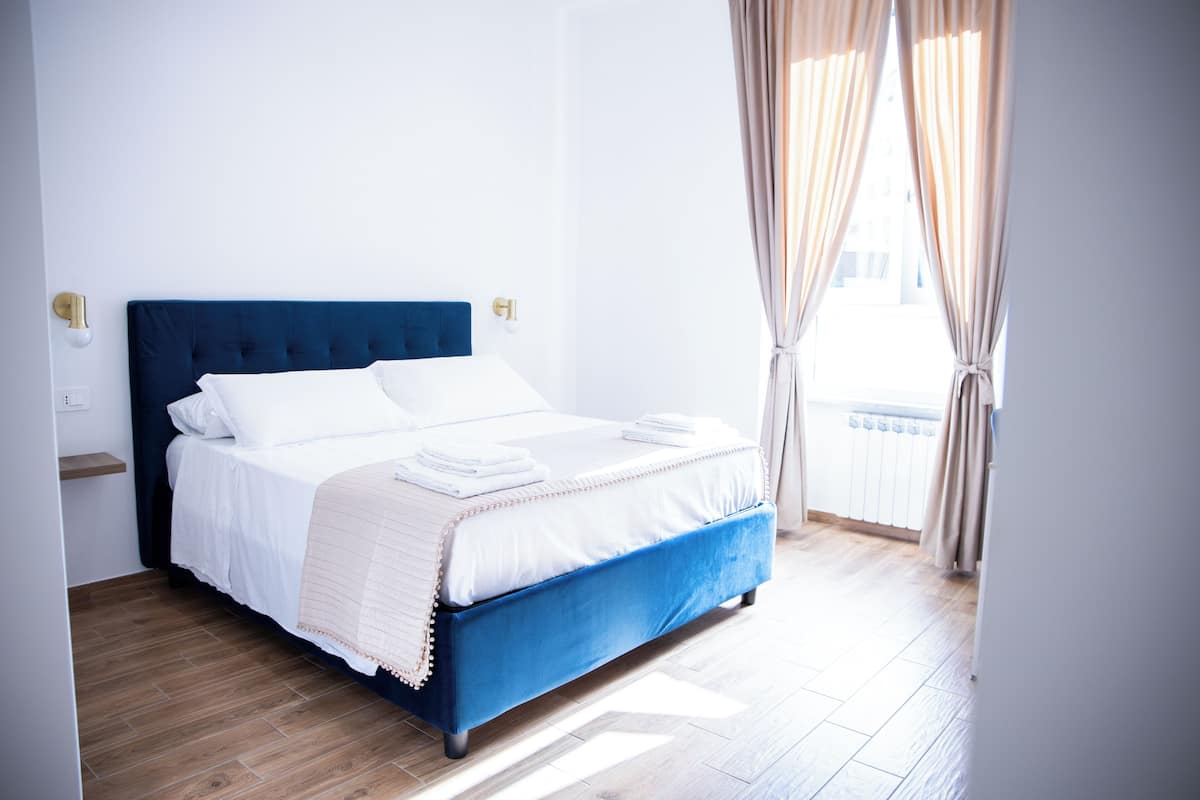Ein ordentlich gemachtes Bett mit weißen Bettwäsche und einem blauen Kopfteil in einem hellen Zimmer mit Holzboden und beigen Vorhängen.
