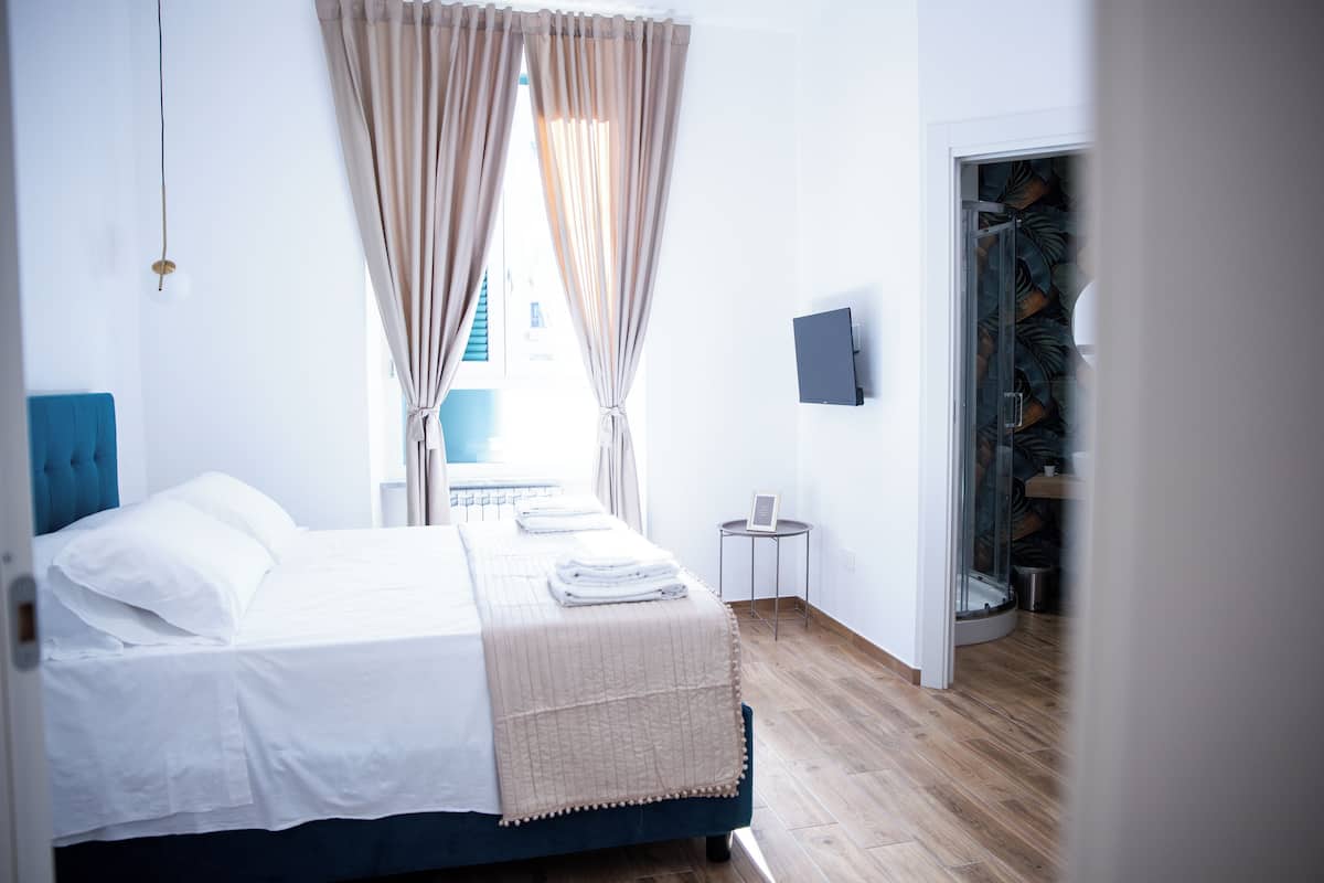 Ein ordentlich bezogenes Bett mit weißen Leinentüchern und einer beige farbenen Tagesdecke, ein kleiner Nachttisch mit einem gerahmten Bild und Blick durch ein Fenster mit beigen Vorhängen nach draußen.