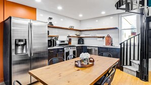 Uma cozinha moderna com uma geladeira de aço inoxidável, forno embutido e máquina de café. Há uma mesa de jantar de madeira com cadeiras e uma bandeja com flores.