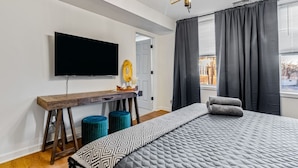 A modern bedroom with a large bed, a flat-screen TV mounted on the wall, a wooden desk, and a window with dark curtains. - Sleeps 12, HOT TUB, Cowboy Pool, Firepits, Yard Games (Louisville)