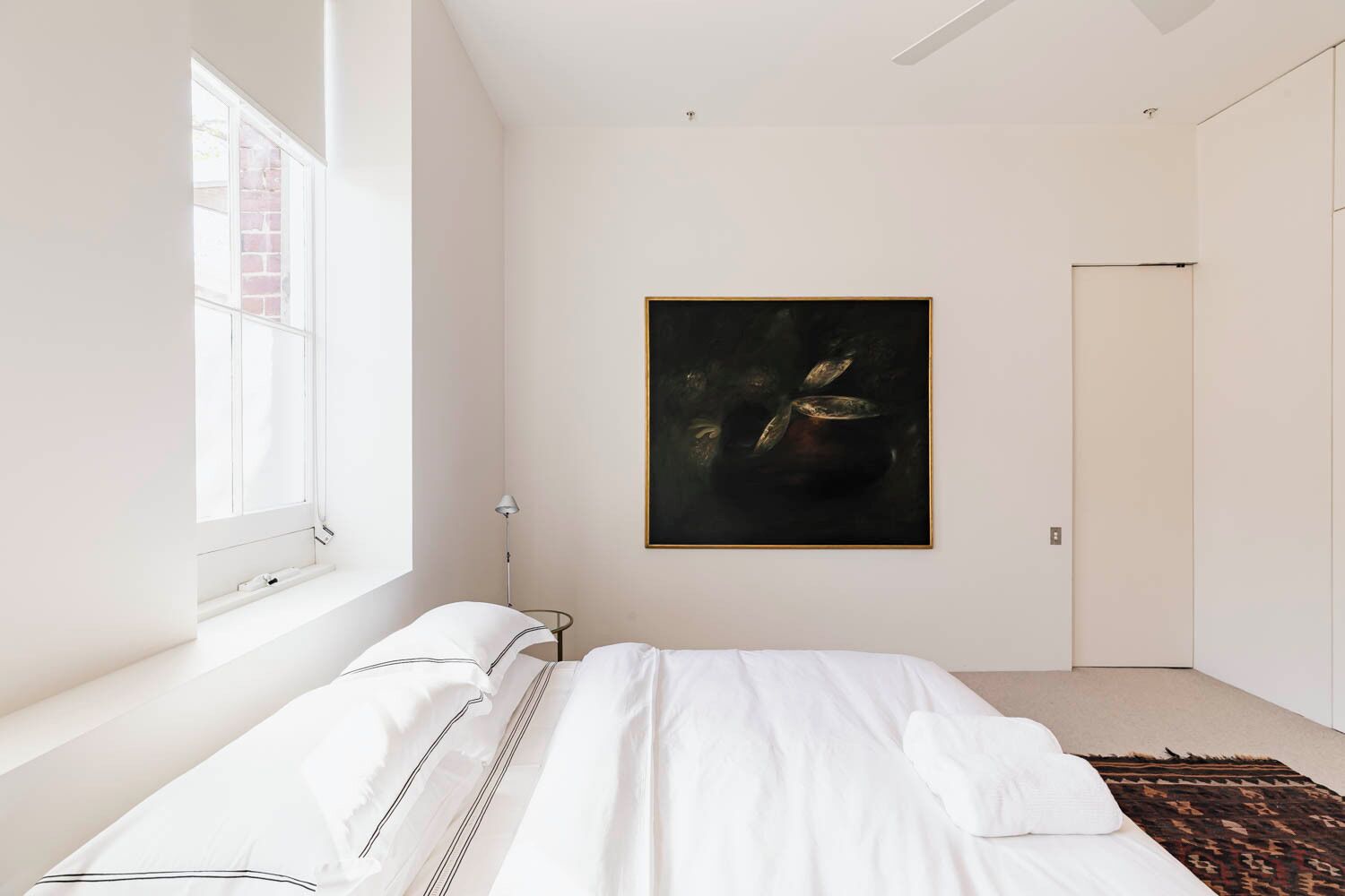 A minimalist bedroom with a large window, a bed with white bedding, and a dark abstract painting on the wall.