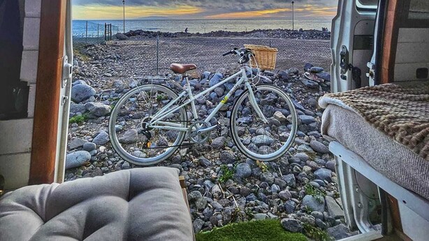 A van interior with a bicycle parked outside, a bed, and a view of a rocky beach at sunset. - Furgot3 1980 (Agulo)