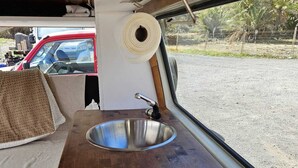 A compact kitchen area with a stainless steel sink, wooden countertop, and a window providing a view of a parking lot and greenery. - Furgot3 1980 (Agulo)