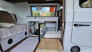 A camper van interior with a bed, a small table, and a window with a view of palm trees. - Furgot3 1980 (Agulo)
