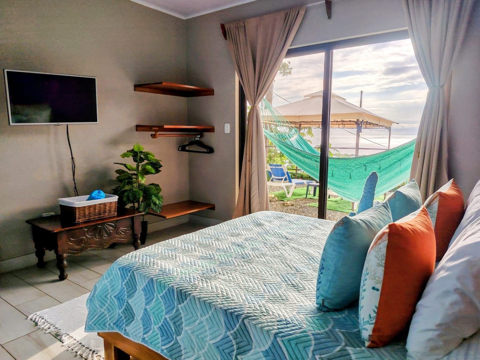 A bedroom with a bed, a TV, a plant, a desk, and a view of the outdoors.