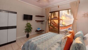 A bedroom with a bed, a TV, a ceiling fan, a plant, and a view of a hammock and sunset. - Mar & Sombra Villas (Quepos)