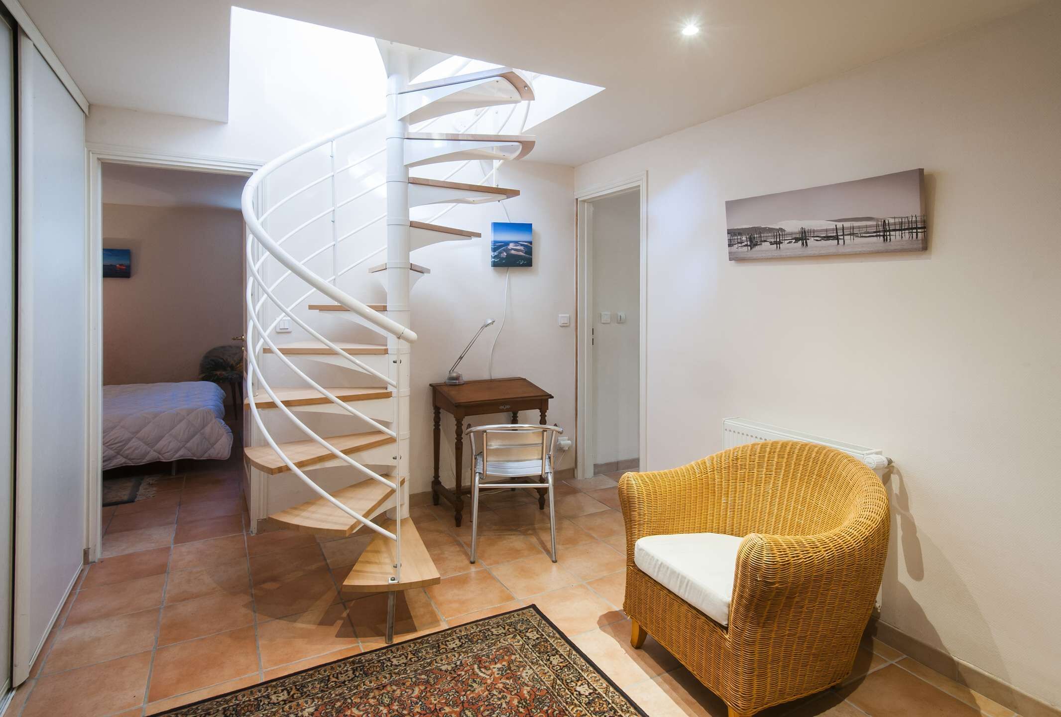 Un escalier en colimaçon menant à une chambre, une chaise en osier, une petite table et un tableau accroché au mur.