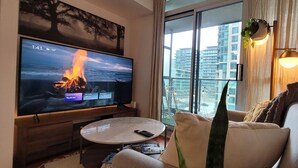 A modern living room with a large flat-screen TV displaying a fire scene, a sofa, a coffee table, and a view of high-rise buildings through a large window. - Studio for 2 in Downtown Toronto  (Toronto)