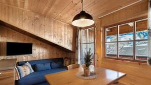 Interior de una cabaña de madera con una mesa de comedor, un sofá azul, un televisor y un ventanal con vistas a un paisaje nevado.