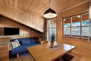 A wooden cabin interior with a dining table, a blue sofa, a television, and a large window with a view of a snowy landscape.