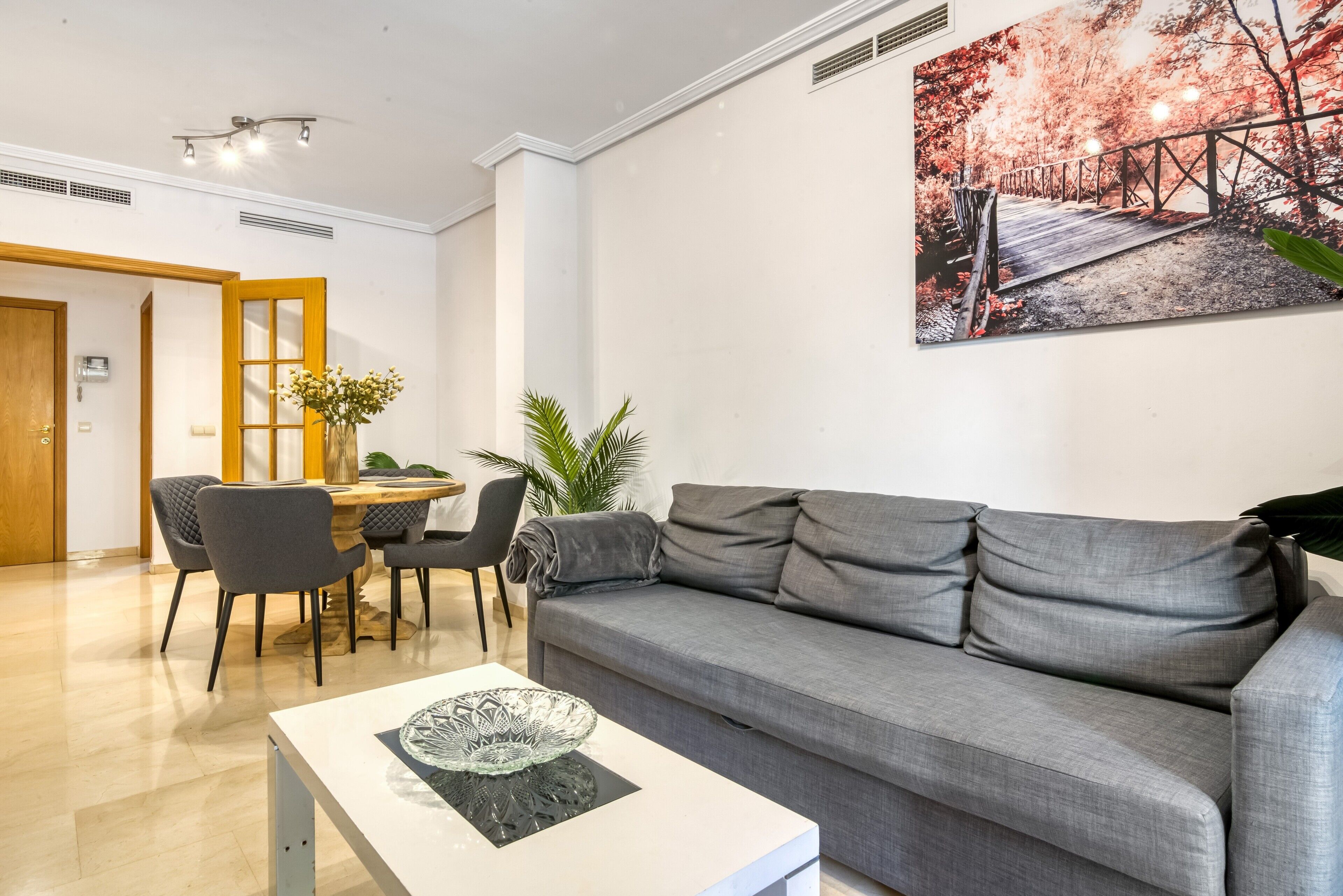 Un salon moderne avec un canapé gris, une table à manger avec des chaises, une table basse surmontée d’un saladier en verre et un grand tableau accroché au mur.