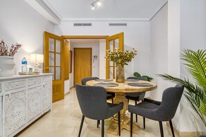 A dining area with a round table, grey upholstered chairs, a white cabinet with intricate carvings, and a vase with flowers.