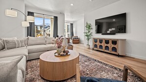 Un salon moderne avec un canapé, une table basse, un téléviseur et une grande fenêtre agrémentée de rideaux.