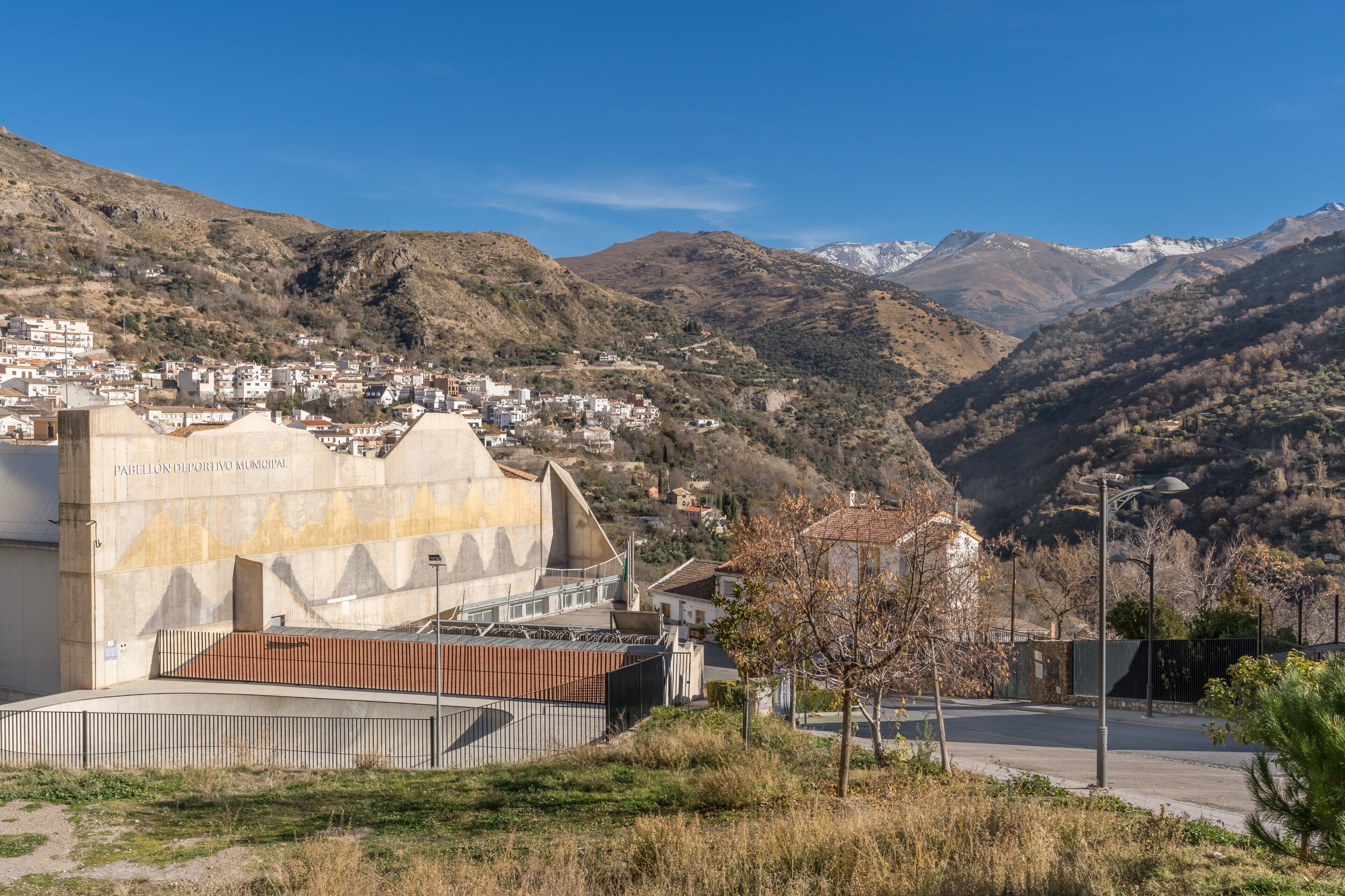 Un paysage montagneux avec un bâtiment portant l’inscription « PARELLON DEPORTIVO MUNICIPAL » et une zone pavée au premier plan.