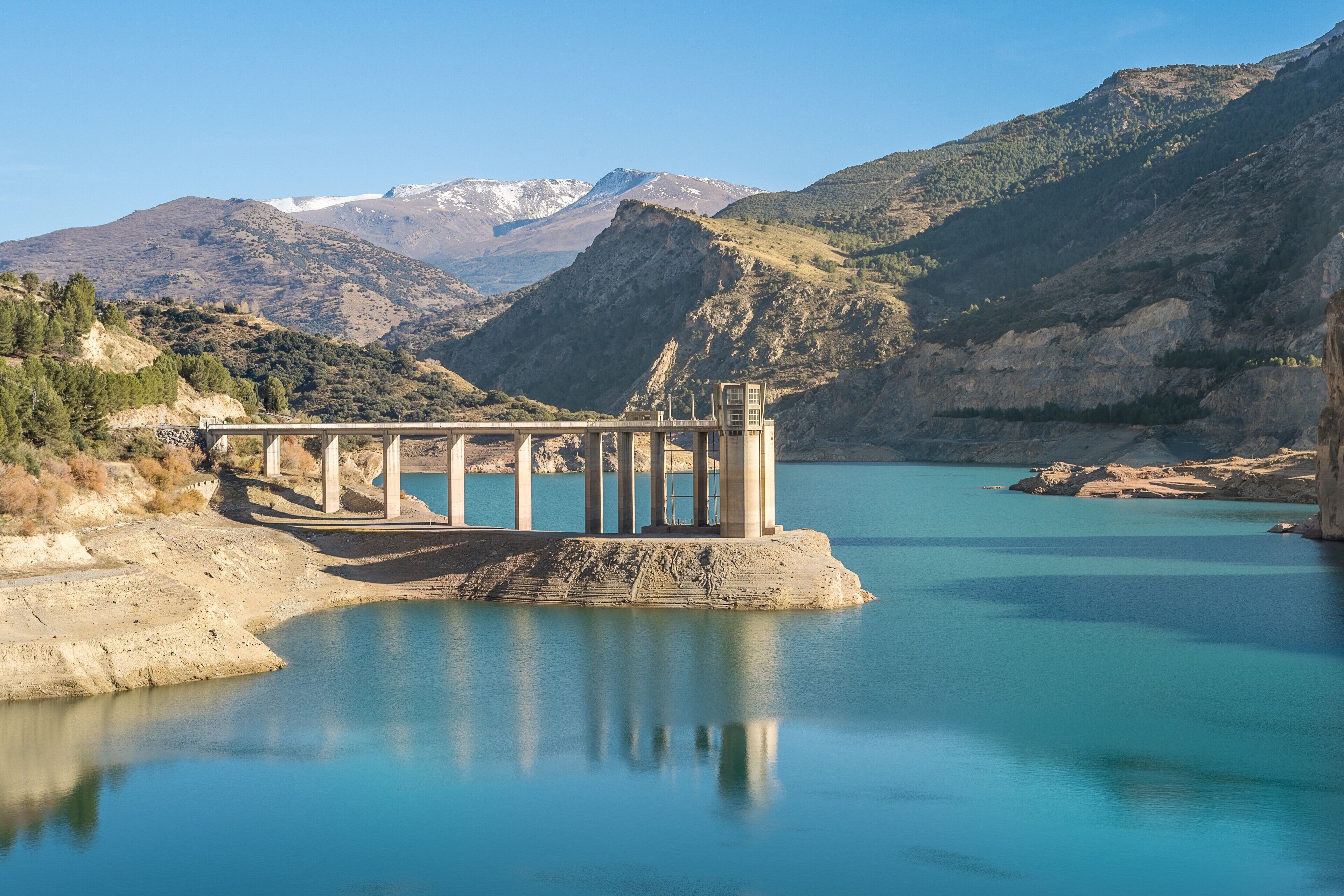 Uma estrutura de barragem com uma ponte, rodeada por montanhas e um lago calmo.