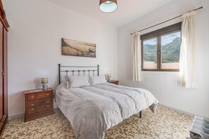 A bedroom with a bed, a nightstand, a lamp, a painting, and a window with curtains.