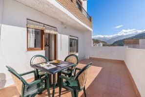 A balcony with a table and chairs, a view of mountains, and a building with white walls and a wooden balcony.
