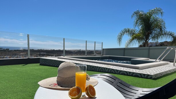 Un espace aménagé au bord de la piscine, avec une pelouse artificielle, un fauteuil de relaxation, une table sur laquelle se trouvent un chapeau et des tranches d’orange, et une piscine dont le bord est recouvert de carreaux.