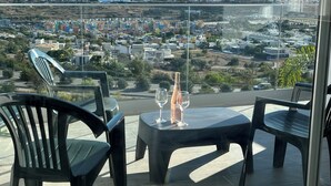 Un balcon avec une table et des chaises, donnant sur une ville côtière avec ses bâtiments et sa végétation.