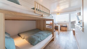 Ein kompakter, moderner Raum mit Etagenbetten, einer kleinen Küchenzeile und Blick durch ein großes Fenster.