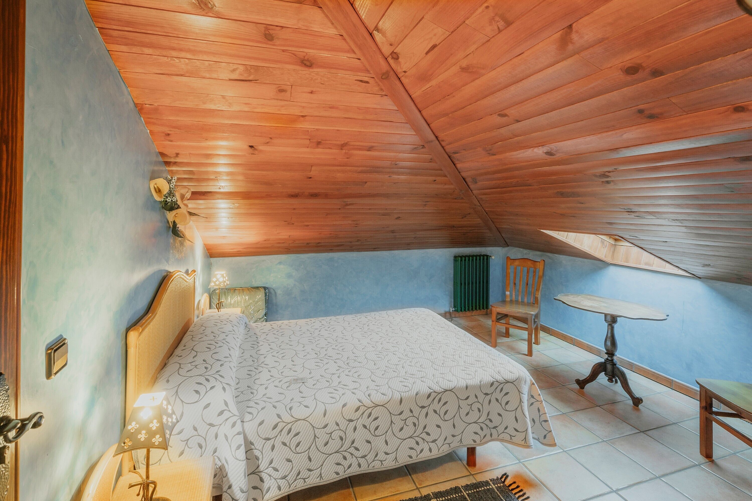 Een slaapkamer met een houten plafond, een bed met een beddengoed met een patroon, een nachtkastje met een lamp, een stoel en een kleine ronde tafel.