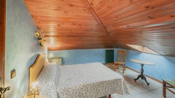 Een slaapkamer met een houten plafond, een bed met een beddengoed met een patroon, een nachtkastje met een lamp, een stoel en een kleine ronde tafel.