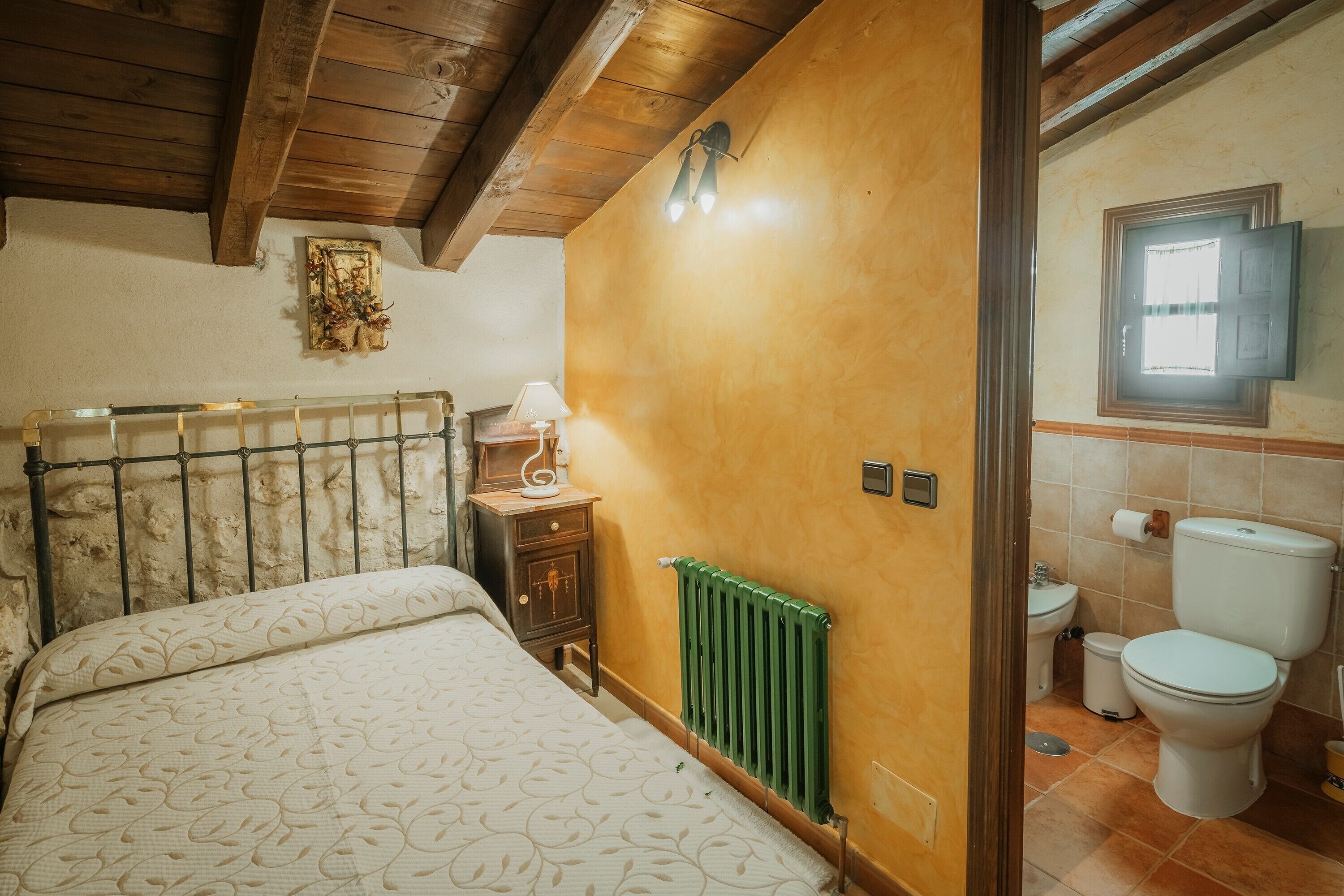 Ein Schlafzimmer mit Bett, Nachttisch, Lampe, Heizkörper und einem Badezimmer mit Toilette und Waschbecken.
