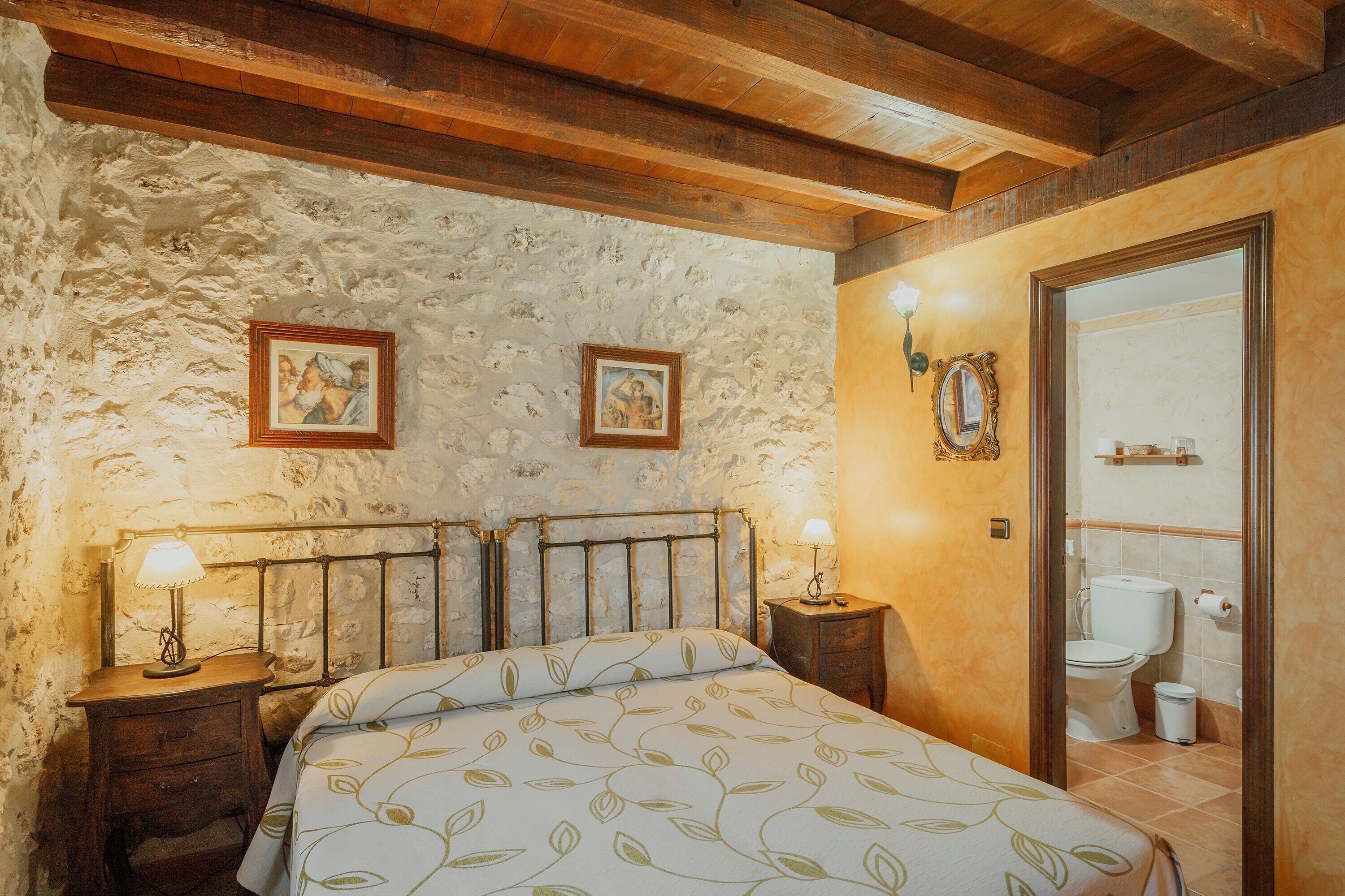 Ein Schlafzimmer mit Steinwand, Holzbalkendecke, einem Bett mit blattgemusterten Bettbezügen, zwei Nachttischen mit Lampen und einem durch eine offene Tür einsehbaren Badezimmer.