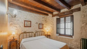 A bedroom with a stone wall, wooden ceiling beams, a bed, two bedside lamps, and a window with shutters.