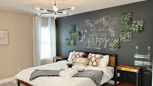 Une chambre à coucher moderne avec un lit, des tables de chevet, un luminaire au plafond et un mur orné d’un texte motivant et de plantes.