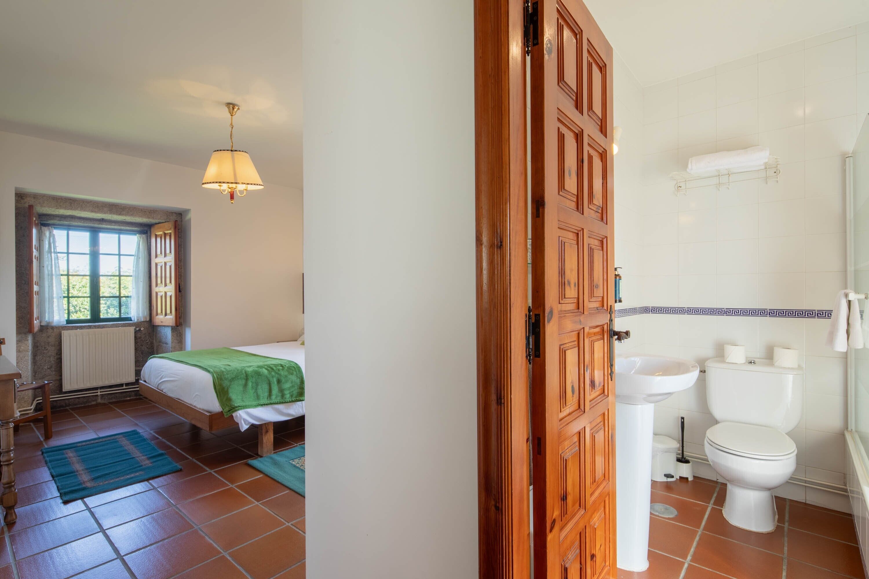 Un dormitorio con cama, ventana, puerta de madera, y baño que incluye inodoro, lavamanos y toallero.
