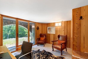 Een kamer met houten wanden, twee leren fauteuils, een zwarte bank en een groot raam met uitzicht op groen.