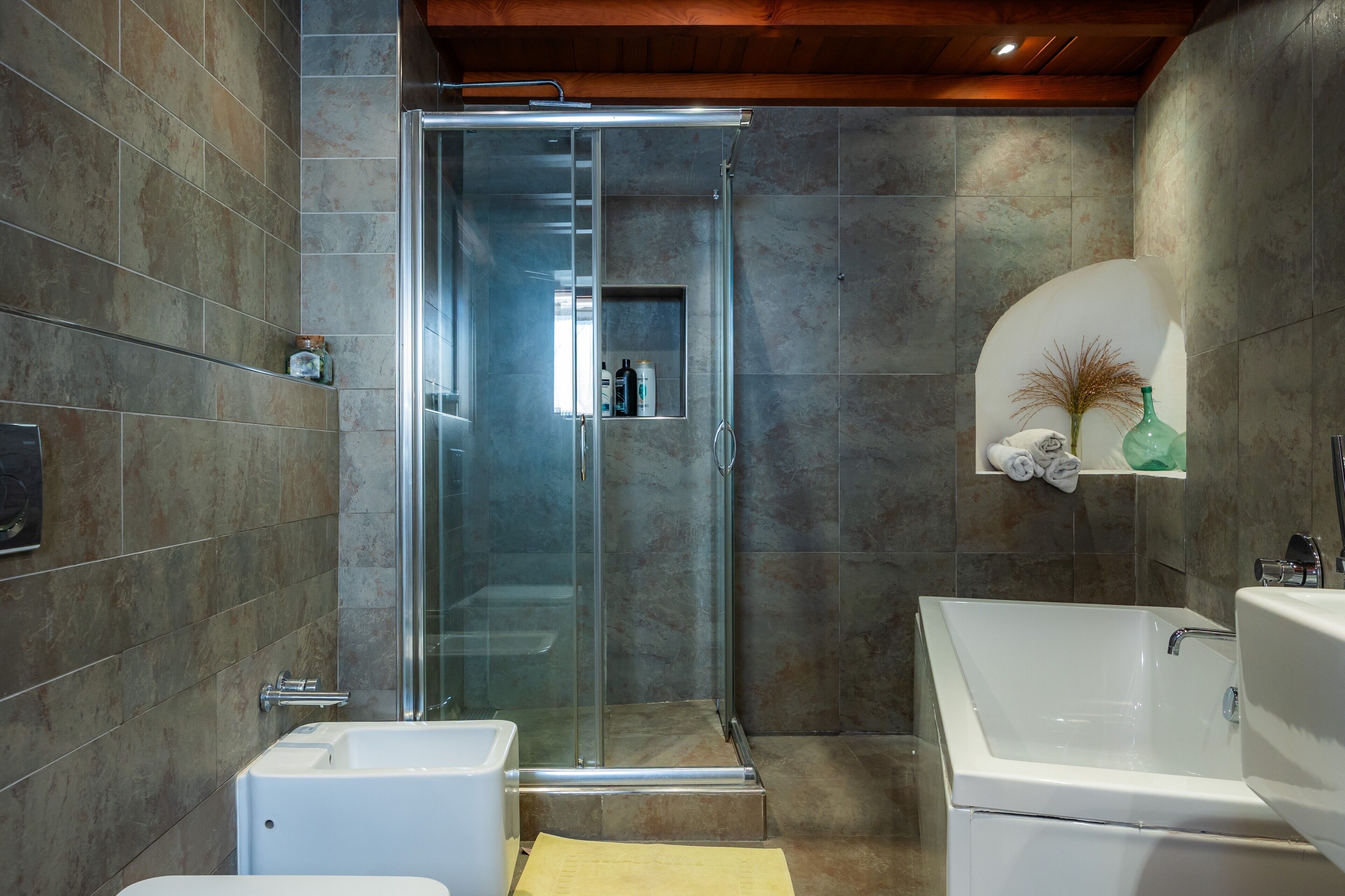 A modern bathroom with a glass-enclosed shower, a white bathtub, and a shelf with toiletries.