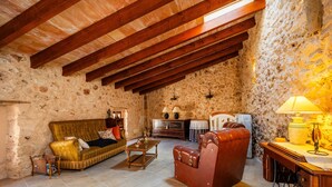 A rustic room with exposed wooden beams, a stone wall, a sofa, a leather armchair, a wooden table, and a lamp.