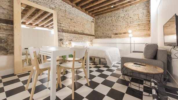 A room with a checkered floor, a bed, a desk with chairs, a small table, and a TV.