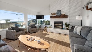A modern living room with a fireplace, comfortable seating, and a large window offering a view. - Ocean Views-Fire Pit-Walk to Beach (Cayucos)