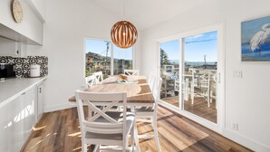 A modern kitchen with a wooden table and chairs, a hanging light fixture, and large windows offering a view of the outdoors. - Ocean Views-Fire Pit-Walk to Beach (Cayucos)