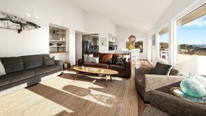A modern living room with a sofa, coffee table, and a view of the kitchen and dining area. - Ocean Views-Fire Pit-Walk to Beach (Cayucos)