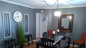 Ein Esszimmer mit Holztisch, Stühlen und einer Chandelier. Zwei Wanduhren und ein Spiegel sind ebenfalls vorhanden.