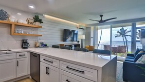 A modern kitchen with white cabinets, a built-in island, and a large window overlooking a scenic view. - Oceanfront, Split AC unit, remodeled November 2024, king bed washer dryer unit (Kihei)