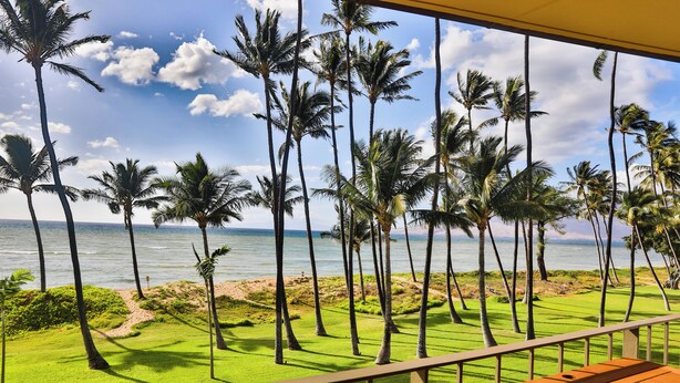 A view from a balcony with a wooden table, overlooking a golf course with palm trees and the ocean. - Oceanfront, Split AC unit, remodeled November 2024, king bed washer dryer unit (Kihei)