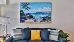 A living room with a blue sofa, a painting of a beach scene, and a lamp on a side table. - Oceanfront, Split AC unit, remodeled November 2024, king bed washer dryer unit (Kihei)