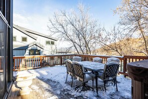 A deck with a table and chairs, surrounded by a wooden railing, with snow on the ground and trees in the background.