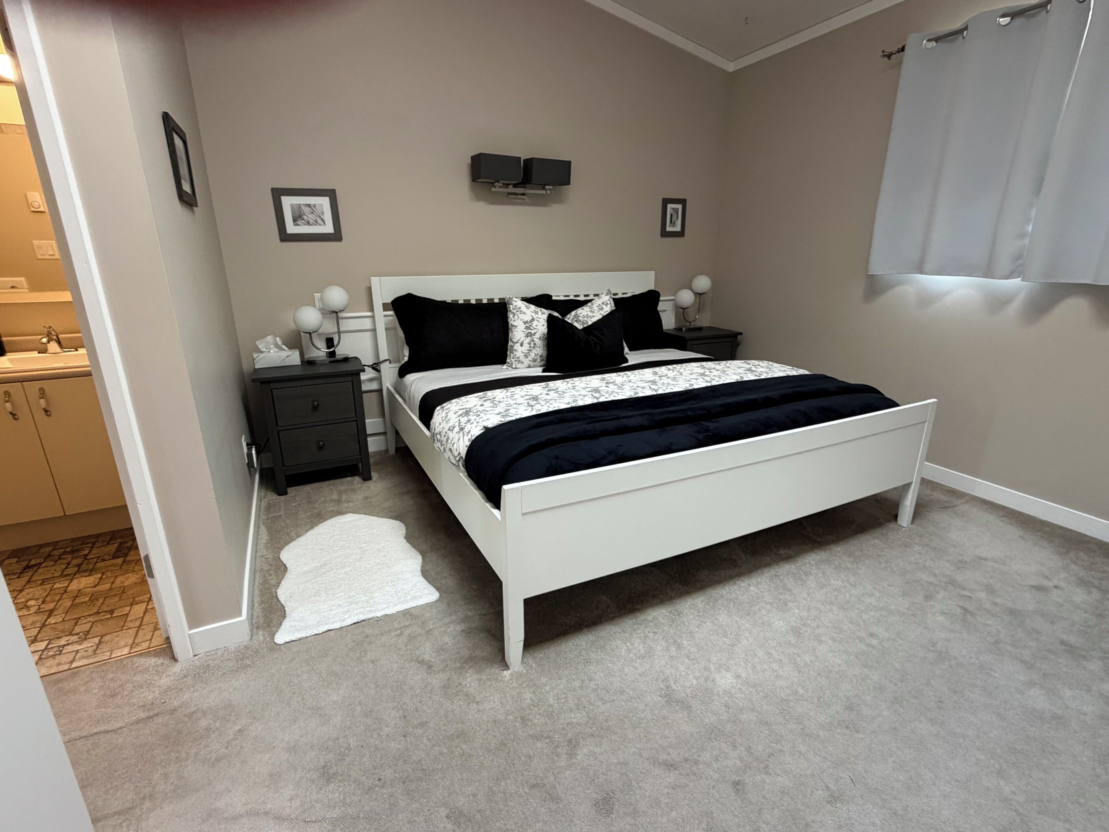 A bedroom with a white bed, a nightstand, a wall-mounted TV, and a bathroom visible through an open door.