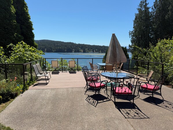 An outdoor patio with a table and chairs, overlooking a body of water and forested hills. - Seascape Suite: Dock, Beach Access, & Free Kayaks! (Sooke)