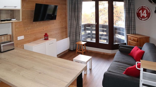 En moderne stue med et træbord, en sofa med røde puder, et fjernsyn monteret på væggen og en balkon med gelænder af træ.
