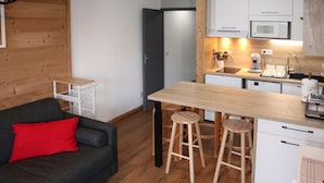 En kompakt opholdsstue med et tekøkken, en sofa med rød pude og et træspisebord med barstole.